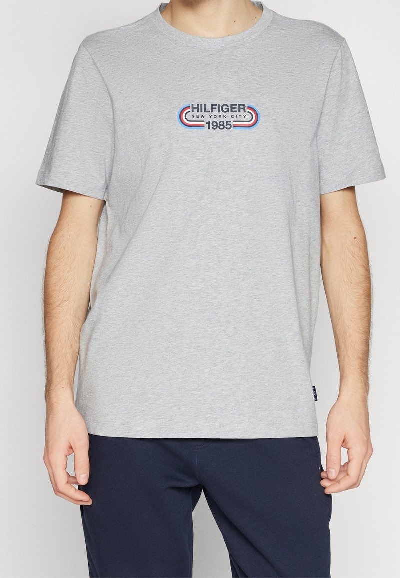 Camiseta de algodón gris de manga corta, que presenta un logo colorido que dice "HILFIGER NEW YORK CITY 1985" en azul, rojo y blanco.