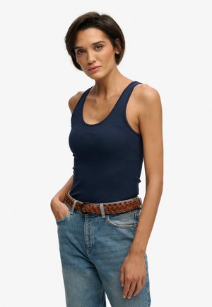 Joven con cabello corto y oscuro, vestida con una blusa sin mangas azul marino, cinturón trenzado marrón y jeans azules, de pie con una mano en el bolsillo.