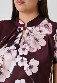 Blusa bordeaux con motivo floreale in tonalità di rosa. Presenta un colletto a chiave e un tessuto morbido dalla consistenza liscia. Maniche corte.