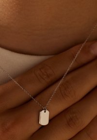 Zilveren ketting met een rechthoekige dog tag-hanger, een glad oppervlak en een kleine lusbevestiging, rustend op een hand.