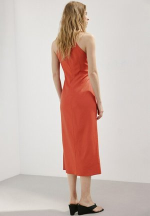 Mujer con cabello largo y ondulado, vistiendo un vestido midi rojo sin mangas y sin espalda, y sandalias negras con tacón, de pie contra una pared blanca lisa.