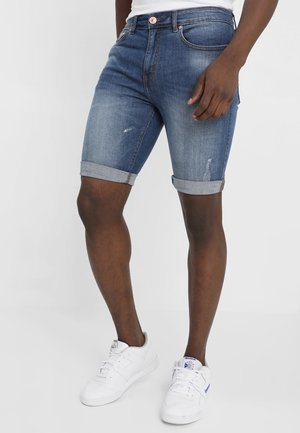 Mand iført opklemte blå denimshorts og hvide sneakers, stående mod en ensfarvet baggrund.