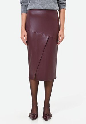 Femme portant une jupe midi en cuir bordeaux avec une fente devant, des collants noirs transparents, des talons pointus bordeaux et un pull gris.