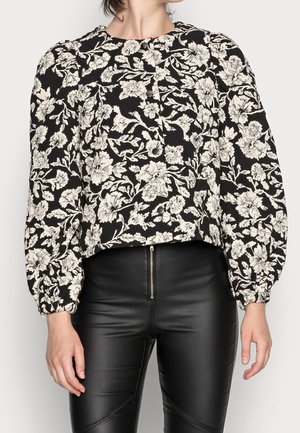 Frau trägt eine schwarz-weiße, geblümte Langarmbluse mit Puffärmeln und schwarze, hoch taillierte Hose mit Reißverschluss vorne.