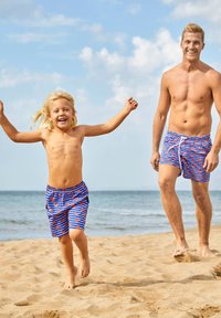 Blaue Badehosen mit roten Herzmustern, getragen von einem Kind und einem Erwachsenen, an einem sandigen Strand, unter einem klaren blauen Himmel.