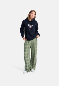 Sweat à capuche marine avec logo et cordons, associé à un pantalon large à carreaux verts. Le modèle porte des baskets blanches. L'arrière-plan est blanc uni.