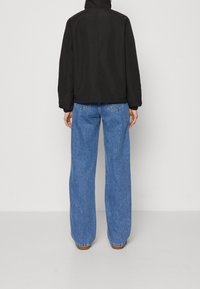 Person klädd i svart jacka och lösa bl å jeans står mot en enfärgad vit bakgrund, vänd bort från kameran.