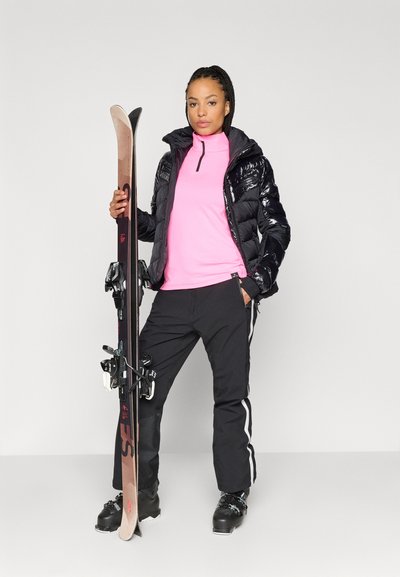 Μαύρο μπουφάν με γυαλιστερό φινίρισμα, ροζ εσωτερικό ρούχο, μαύρο παντελόνι σκι με λευκές ρίγες, κρατώντας ξύλινες σανίδες σκι με ιμάντες. Μαύρα μποτάκια σκι.
