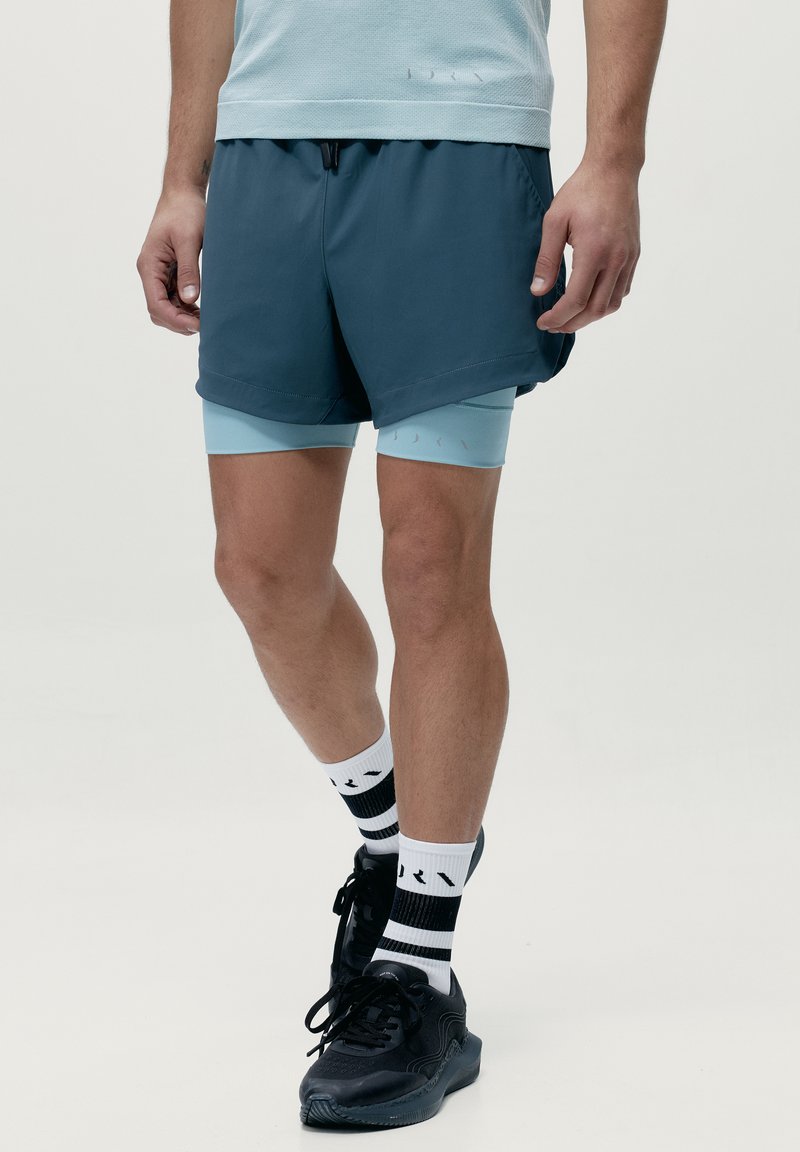 Herrarnas atletiska outfit med mörkblåte shorts över ljusblå kompressionsshorts, i kombination med svarta sneakers och randiga svartvita strumpor.