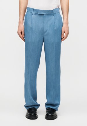 Mees kannab helesiniseid voltidega pükse ja musti ametlikke kingi, seisab ühevärvilise taustaga.