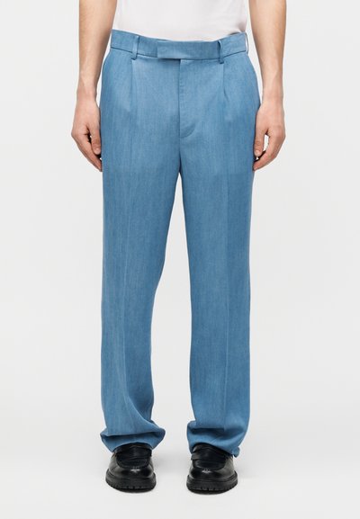 Homme portant un pantalon plissé bleu clair et des chaussures de ville noires, debout devant un fond uni.