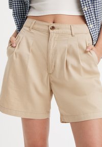 Shorts plissés beiges en tissu léger, dotés d'une fermeture à bouton sur le devant et de poches latérales, avec un ourlet soigné et une texture lisse.