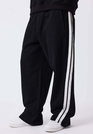 Pantalon noir ample avec deux bandes blanches sur les côtés, porté avec des baskets blanches sur un fond uni.