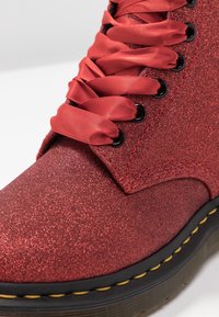 Röd glittrig ankelboot med glänsande satinsnören, svarta ögla och en tjock svart sula med gula sömnadsdetaljer. Närbild på övre delen.