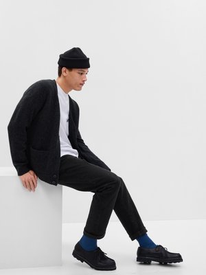 Dark gray knitted cardigan with front pockets, worn over a white t-shirt. Black pants, blue socks, and black shoes, seated on a white surface.