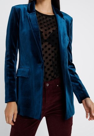 Vrouw die een teal fluwelen blazer draagt over een zwart doorschijnend polkadot-topje en donkerrode broek, staand tegen een effen achtergrond.