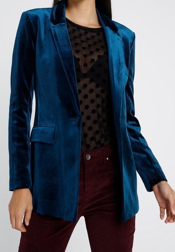 Femme portant un blazer en velours bleu sarcelle sur un haut noir transparent à pois et un pantalon rouge foncé, debout devant un fond uni.