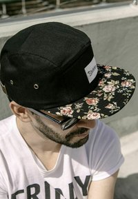 Black cap with a flat floral-patterned brim. Features a white label on front. Circular metal eyelets on the sides for ventilation.