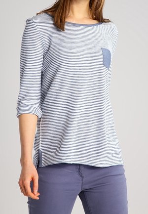Femme portant un haut à manches longues rayé bleu et blanc avec une poche poitrine bleue et un pantalon bleu, debout avec le bras gauche détendu.