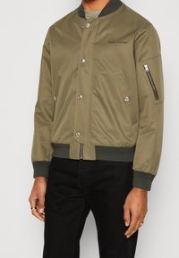 Oliivinvihreä bomber-takki, jossa on vetoketjullinen hiha-tasku, nepparikiinnitys ja resorit hihansuissa. Takissa on sileä, kevyt kangas ja istuva malli.