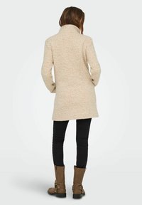 Manteau en mélange de laine beige avec un col haut, surface texturée, coupe mi-longue, associé à un jean slim noir et des bottes marron jusqu'aux genoux.