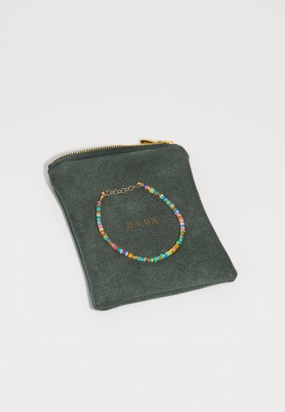Une pochette zippée verte avec un collier de perles colorées exposé dessus, portant le texte "HA HA" estampillé en doré.