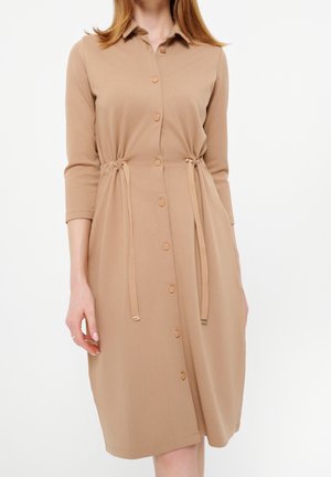 Femme portant une robe beige à manches longues, boutonnée, avec col et ceintures ajustables à la taille, debout devant un fond blanc uni.