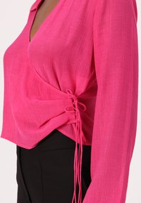 Pink blouse with a textured finish, featuring a deep V-neck, long sleeves, and ruched detail on the side with lace-up ties. Paired with black pants.