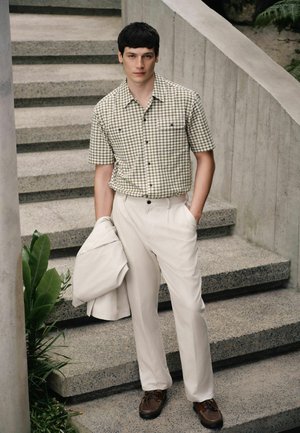 Bărbat purtând o cămașă cu mânecă scurtă, cu model, și pantaloni crem ține o jachetă crem, stând pe scări din beton lângă plante verzi.