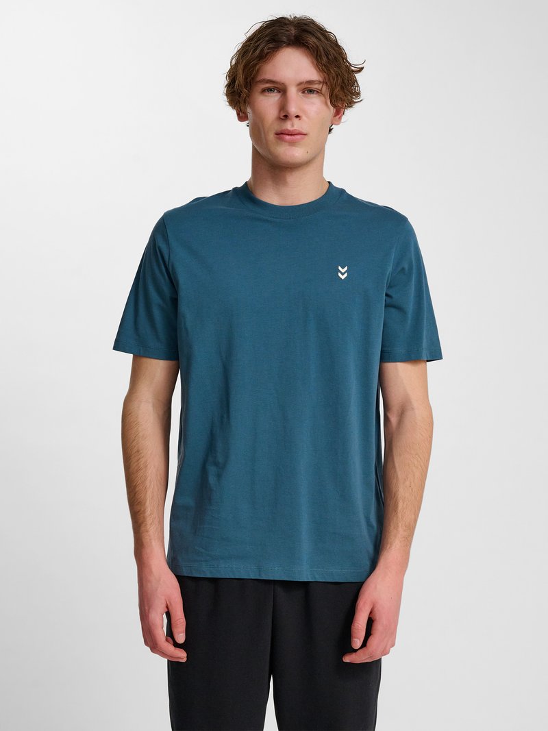 Junger Mann mit kurzen lockigen Haaren, der ein blaues T-Shirt mit kleinem weißem Logo auf der linken Brust trägt und vor einem weißen Hintergrund steht.