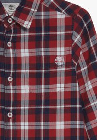 Koszula w kratę w kolorach czerwonym, białym i granatowym, zapinana na guziki, z kieszenią na piersi i wyszywanym logo. Posiada kontrastowe guziki i teksturowaną tkaninę.