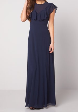 Femme portant une robe maxi bleu marine sans manches avec un détail volanté sur la poitrine et un tissu transparent à l'encolure et aux épaules.