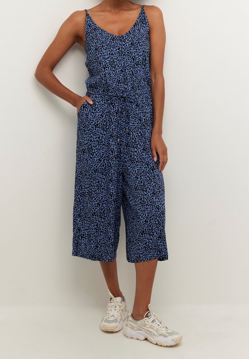 Femme portant une combinaison sans manches bleue et noire avec une taille à cordon et des baskets blanches, se tenant devant un mur clair.