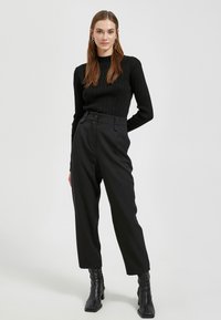 Czarny, prążkowany top z długim rękawem w połączeniu z czarnymi spodniami typu high-waist. Modelka ma na sobie czarne botki za kostkę, co podkreśla elegancki, dopasowany wygląd.