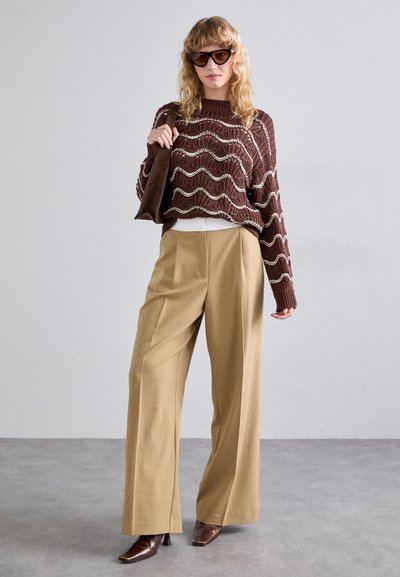 Brun strikket sweater med hvide bølge mønstre, parret med beige vide ben bukser og brune spidse hæle. Accessories inkluderer mørke solbriller.