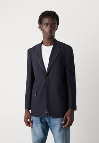 Blazer azul-marinho com costura branca contrastante, botão frontal único, lapela entalhada, dois bolsos frontais, combinado com uma t-shirt branca e jeans azuis.
