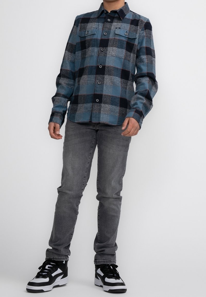 Chemise en flanelle à carreaux bleus, noirs et gris avec deux poches poitrine ; associée à un jean slim gris et des baskets noir et blanc.