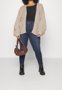Beige gebreid vest met losse mouwen, zwart geribd topje, donkerblauwe jeans en een bruine leren handtas, gecombineerd met zwarte enkellaarsjes.