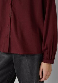 Burgundy long-sleeve shirt with a button-down front, featuring a relaxed fit and gathered cuffs. Paired with dark gray jeans.