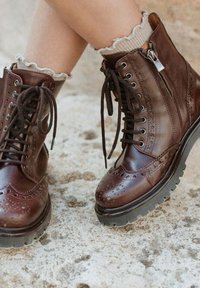Botas de cuero marrón hasta el tobillo con detalles de ala, punta redonda, cierre de cordones al frente, cremallera lateral y suela gruesa de goma negra. Superficie texturada.