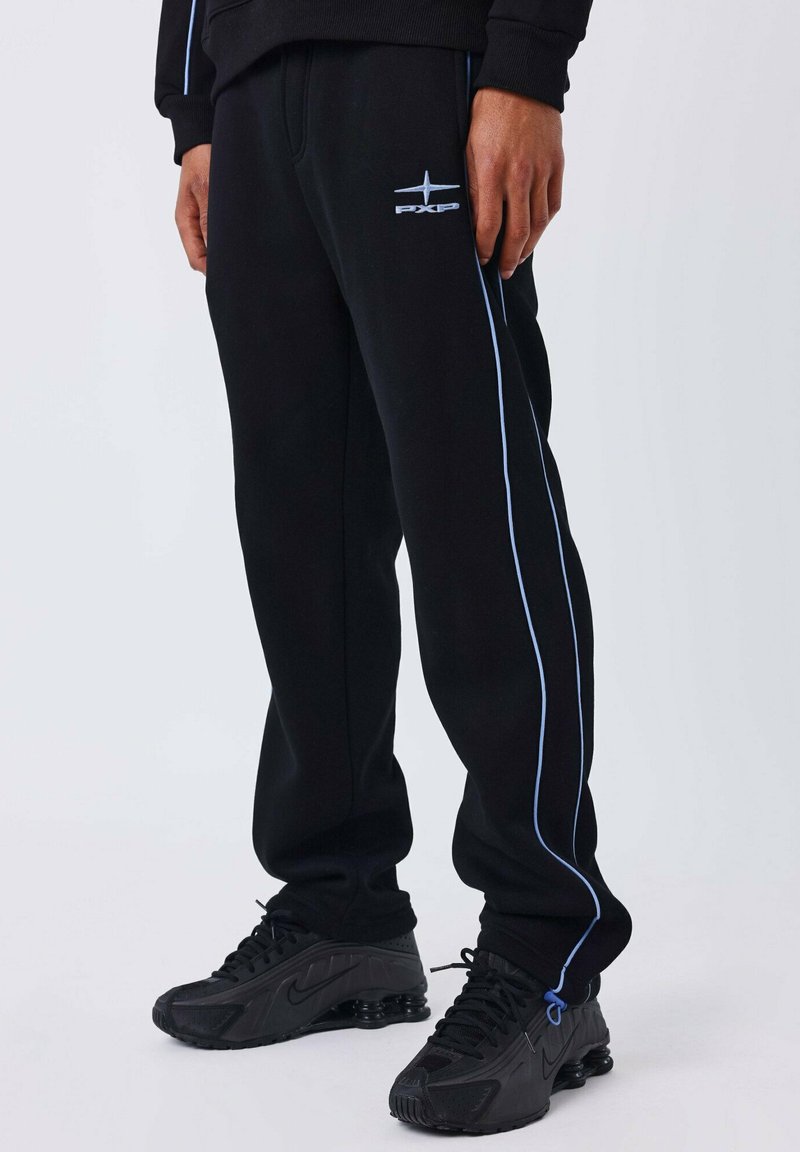 Persona que lleva pantalones de chándal negros con franjas laterales azules y un logo bordado, combinados con zapatillas Nike negras.