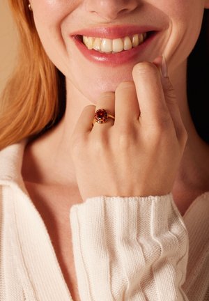 Goldring mit einem großen roten ovalen Stein, umgeben von kleinen bunten Steinen, getragen an einer Hand mit einem leicht strukturierten Strickpullover.