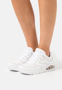 Witte sportieve schoenen met een gestructureerde mesh-bovenkant, tan accenten en een zichtbare luchtkussen in de zool. Beschikt over een ronde neus en vetersluiting.
