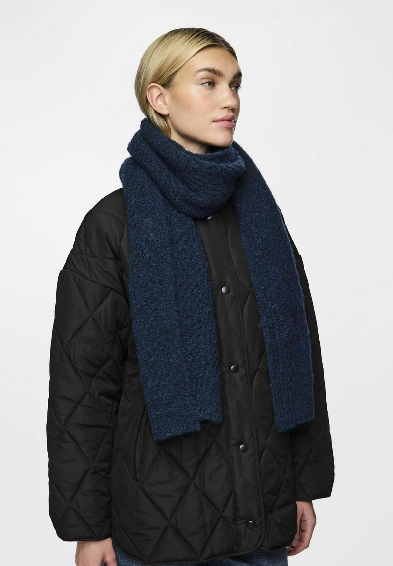 Écharpe en laine bleu marine avec une finition texturée, enroulée autour du cou, portée avec une veste noire matelassée à boutons-pression.