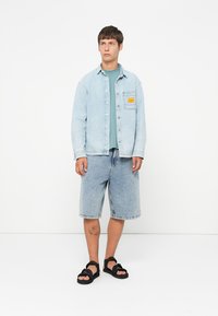 Young man stands wearing a light denim jacket over a green t-shirt, knee-length denim shorts, and black strapped sandals on a white background.