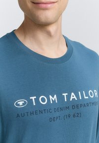 Modrá bavlnená tričko s okrúhlym výstrihom, na ktorom je biely a tmavomodrý text "TOM TAILOR" a "AUTHENTIC DENIM DEPARTMENT".