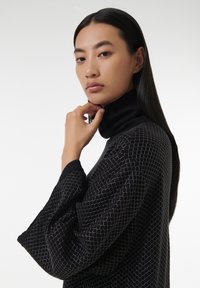 Un cuello alto negro con un patrón de rejilla geométrica en plata, que presenta mangas amplias y un cuello ajustado, usado por una persona con cabello largo.