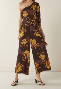 Mono floral marrón con un diseño de un solo hombro, pierna ancha y decorado con flores amarillas, acompañado de un cinturón a juego.