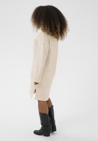 Pleteni pulover haljina u svijetlosmeškoj boji s dugim rukavima, bočnim prorezima i opuštenim krojem, uparen s crnim kožnim čizmama do koljena.