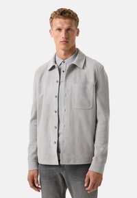 Grijs suède jasje met een kraag, voorzak en knoopsluiting, gecombineerd met een lichtgrijs shirt eronder. Zachte textuur.